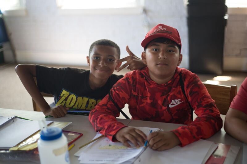The image shows two young boys sitting at a table. The boy on the left is wearing a black t-shirt with the word "ZOMBIE" on it. The boy on the right is wearing a red cap and a red shirt. They both appear to be looking at the camera. There are papers and a water bottle on the table.